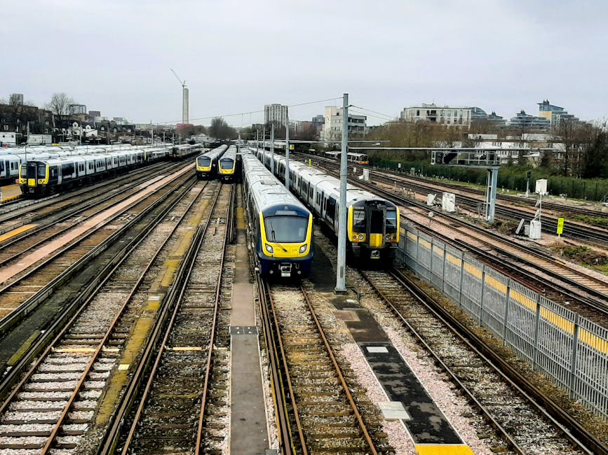 The image depicts a modern train station platform with a red and black train stopped at the platform, which is clean and well-maintained. The platform surface is smooth with tactile paving strips alon